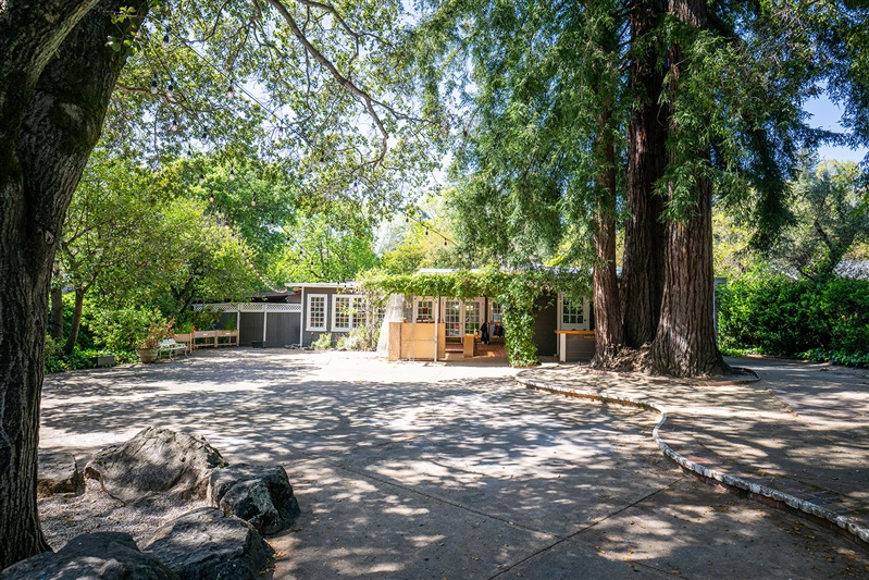 A picture of a one-story building from across a paved patio, shaded by trees.